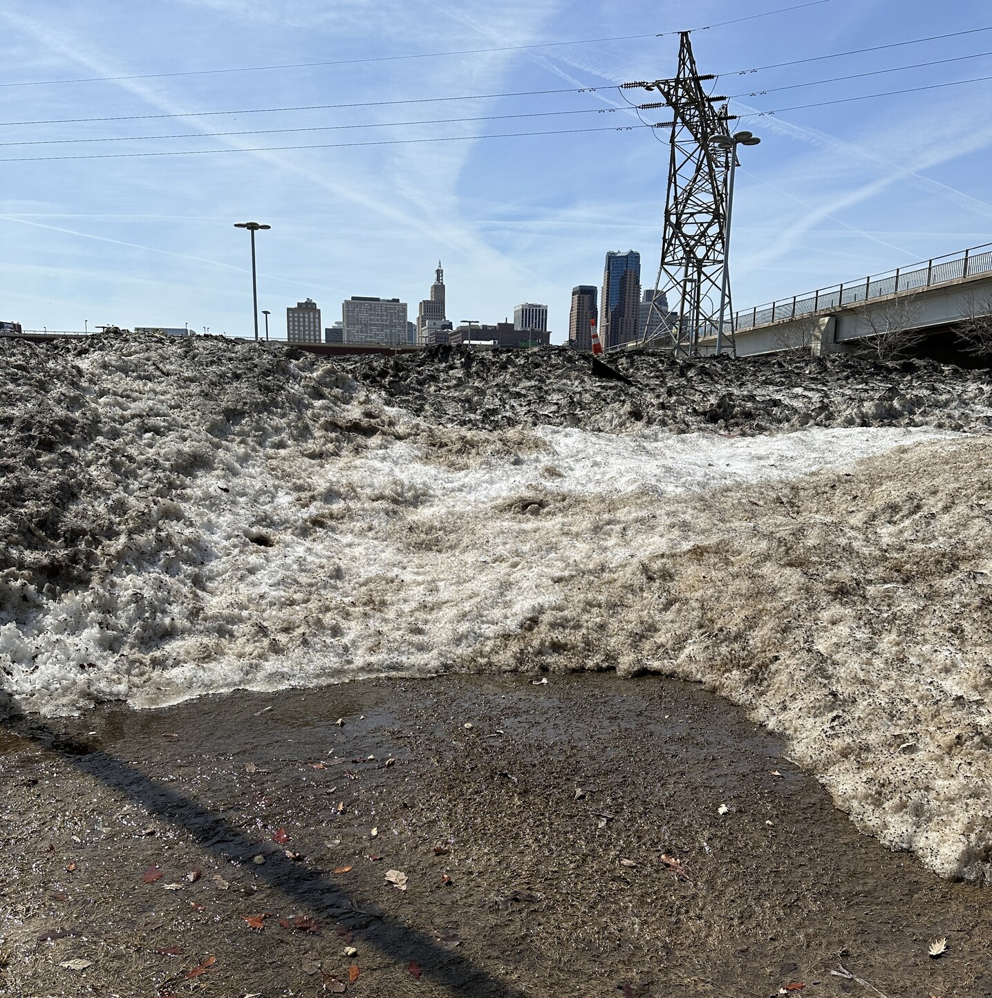 Large pile of “dirty” snow in downtown Saint Paul’s Union Depot Lot C in the sunshine with a pool of water in front and the downtown skyline in the back
