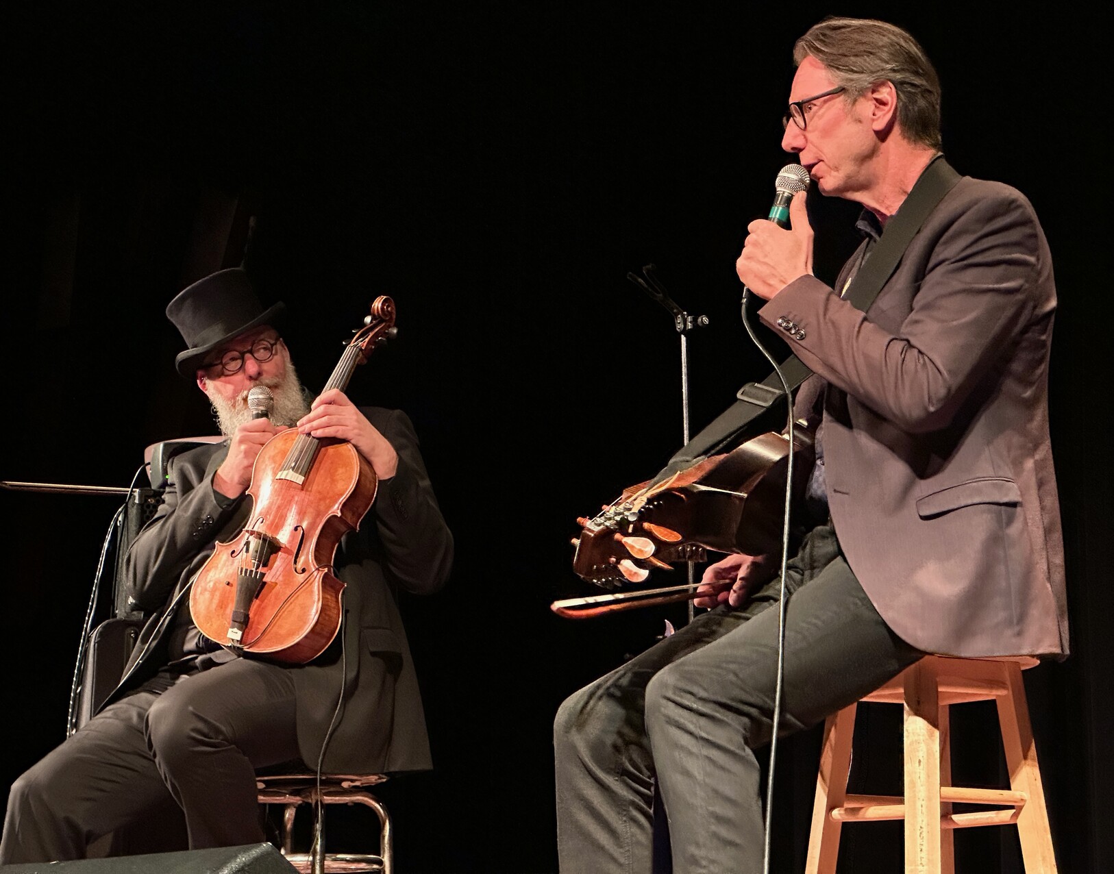 Mikael Marin in a top hat holding a baby cello while Olov Johansson holds his nyckelharpa in one hand and a microphone in the other - both are sitting on stools on stage at the Cedar Cultural Center