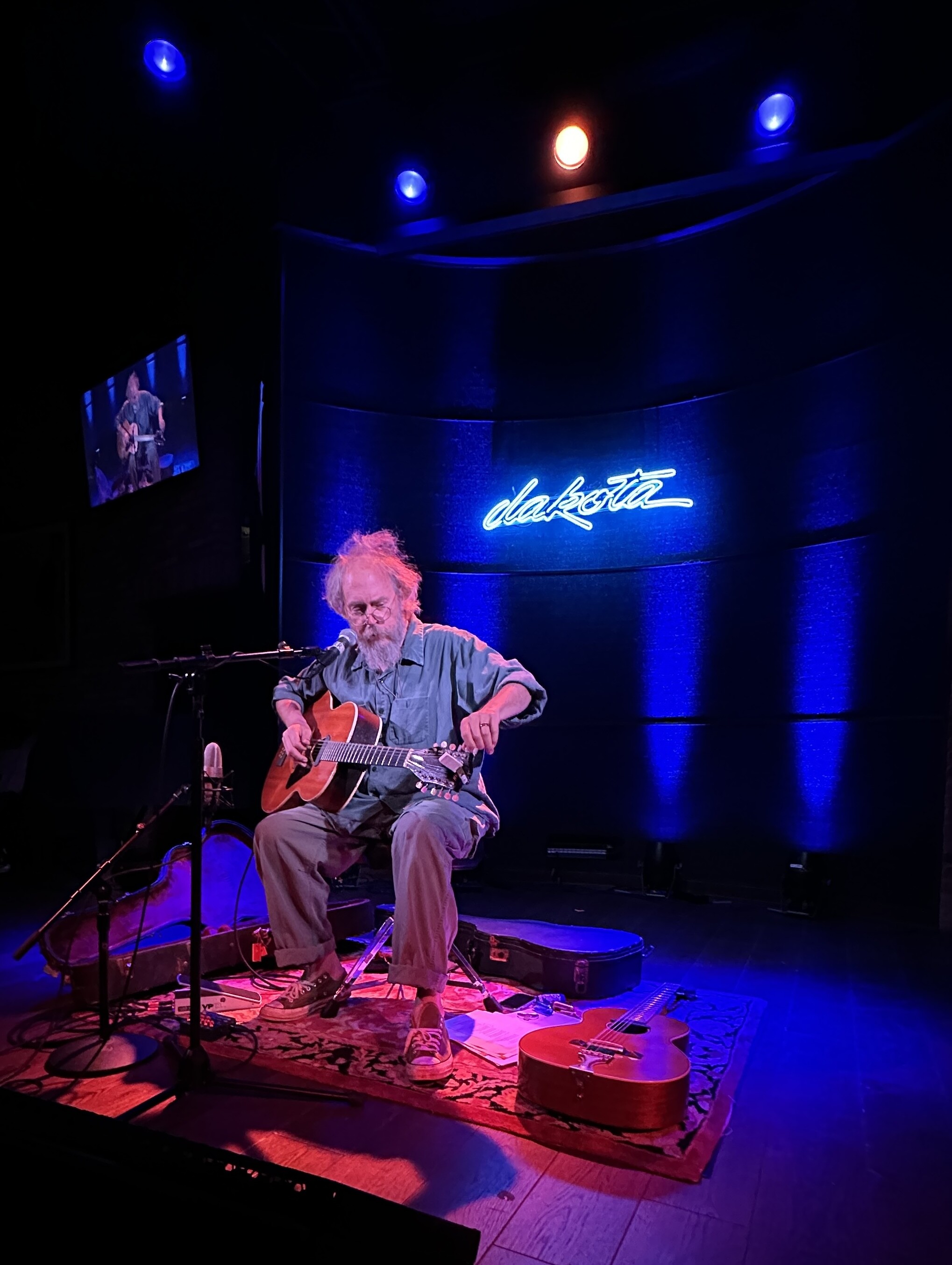 Charlie Parr sitting on stage in red and blue spotlights holding a guitar with a blue neon “dakota” sign behind him