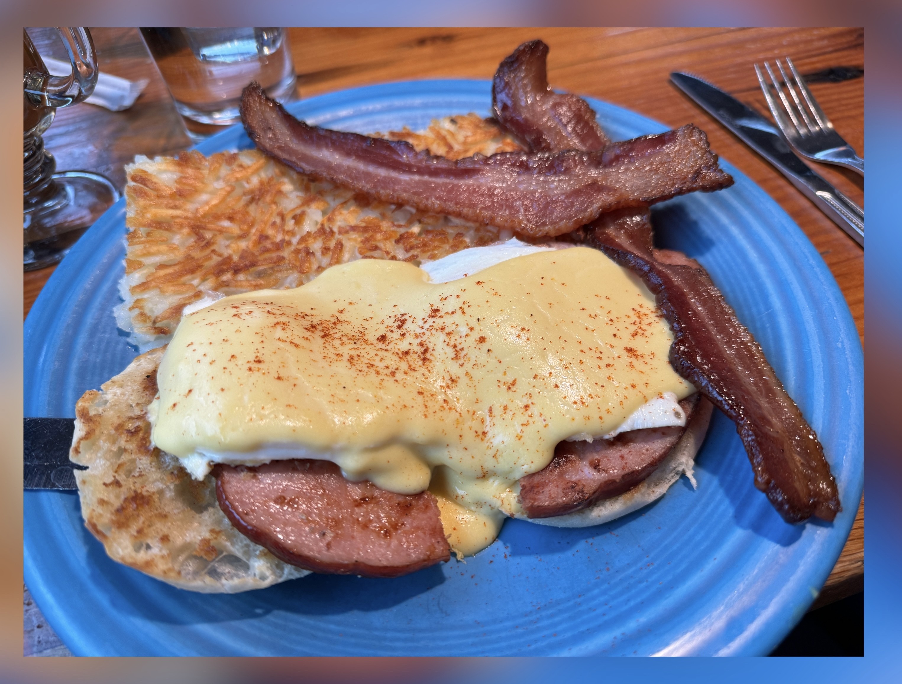 Eggs Benedict on a blue plate with hash browns and two strips of bacon