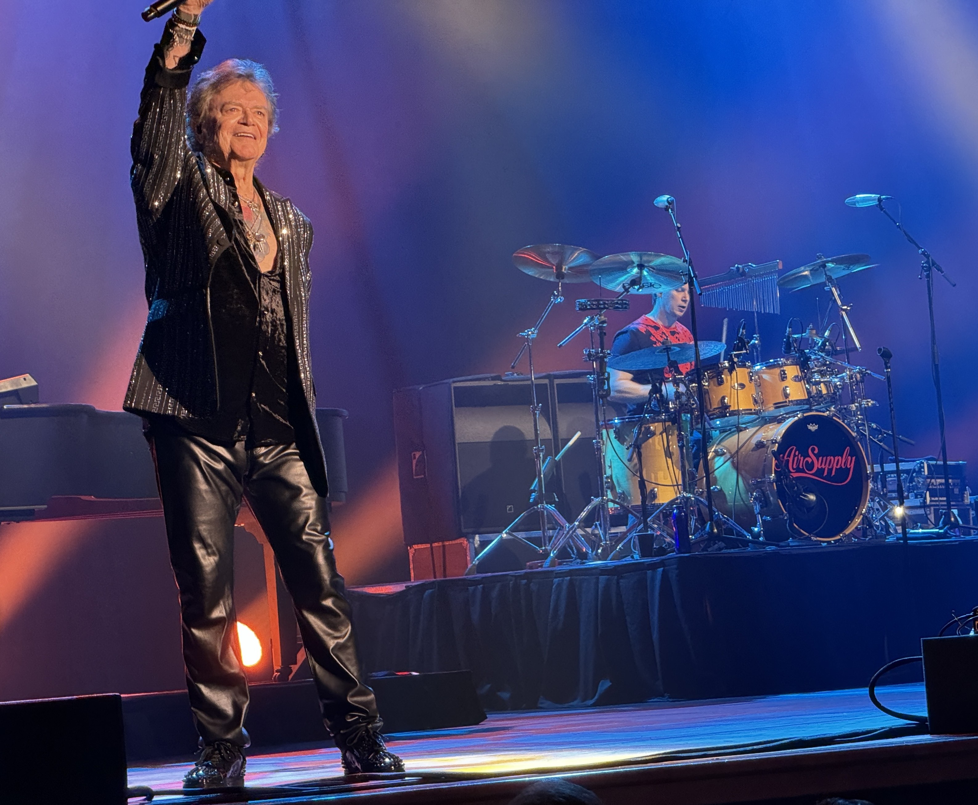 A smiling Russell Hitchcock on stage at Mystic Lake Casino with a raised mic, black leather pants and a sparkling black sequin jacket with a drummer in the background