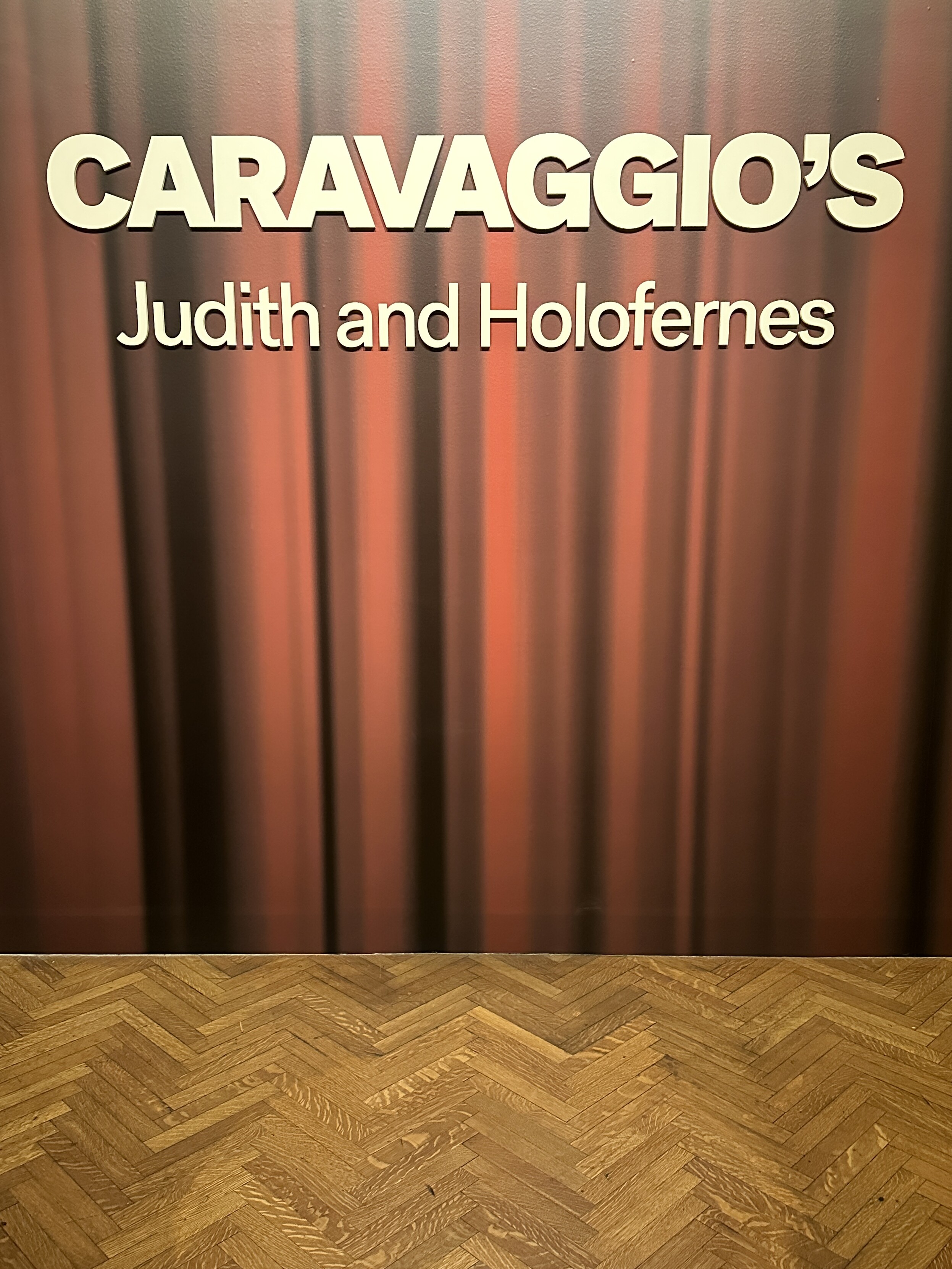 Caravaggio’s Judith and Holofernes signage at the Minneapolis Institute of Art, with the words in white against a red curtain backdrop and zig-zag wood floor pattern below