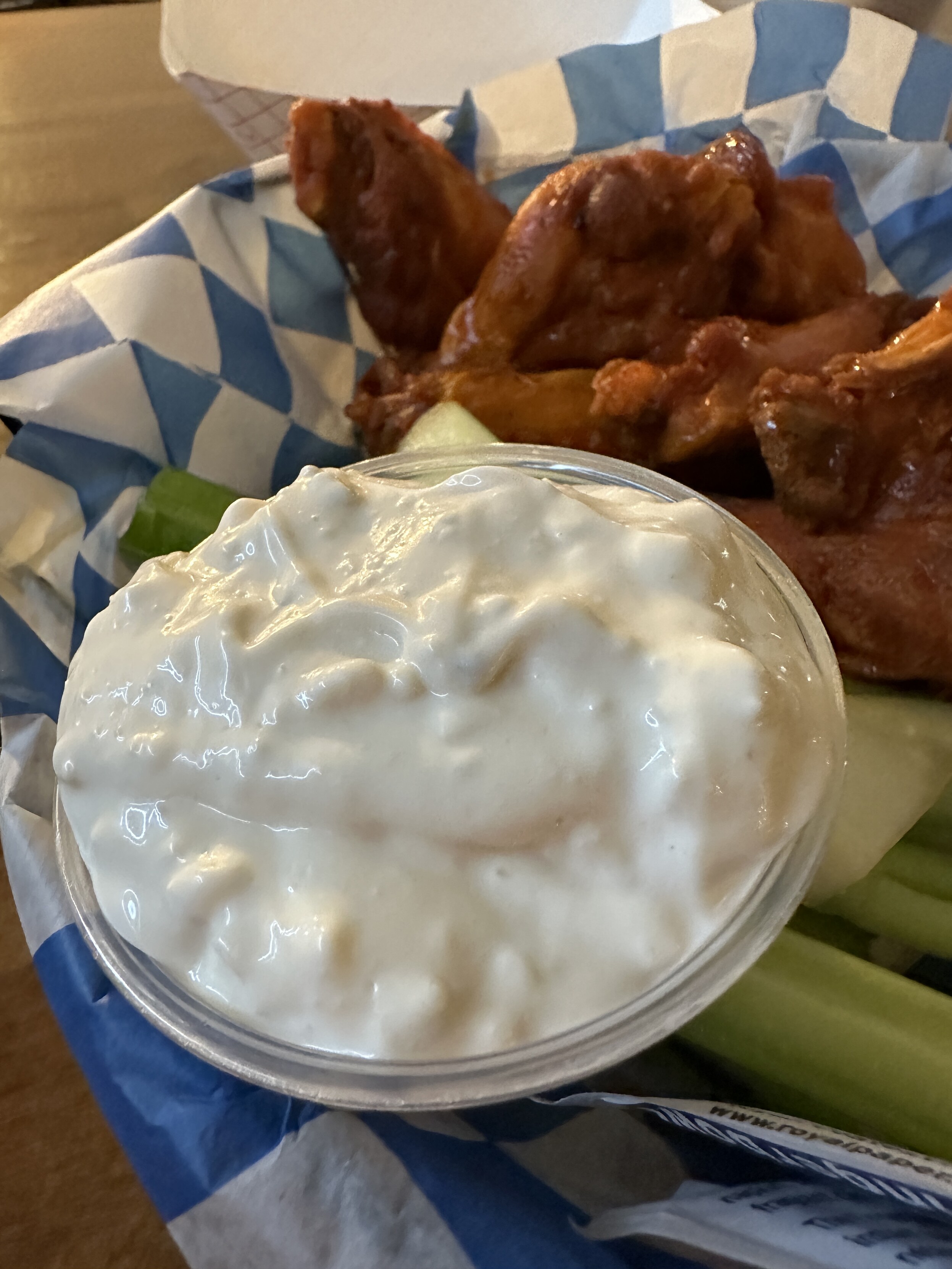 Giant cup of homemade blue cheese sitting in a basket of chicken wings and celery with a blue and white checkered paper liner