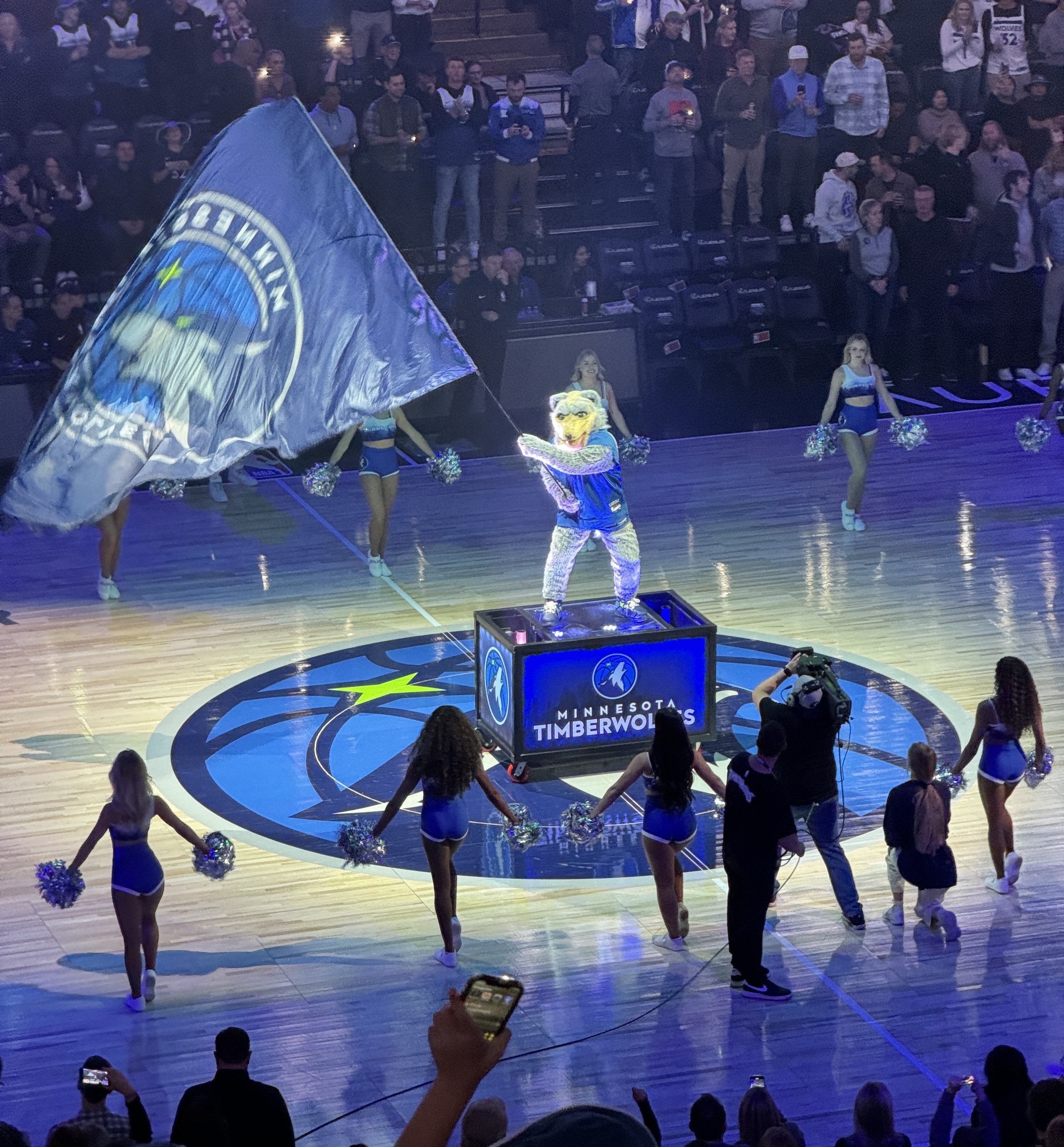 Timberwolves mascot Crunch waving a giant flag at center court, surrounded by cheerleaders 
