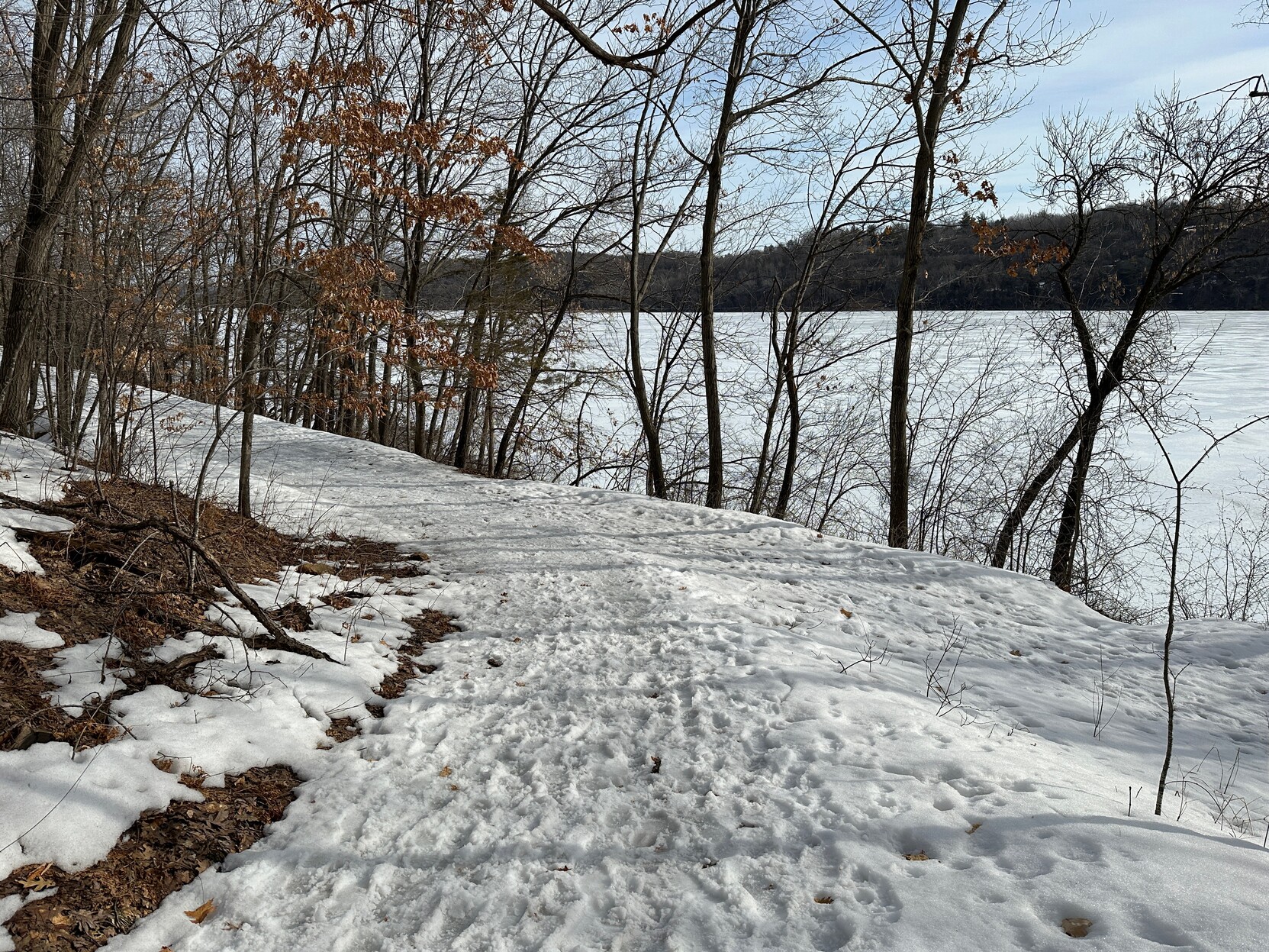 Snow and ice-covered riverside trail with fall leaves mixed in