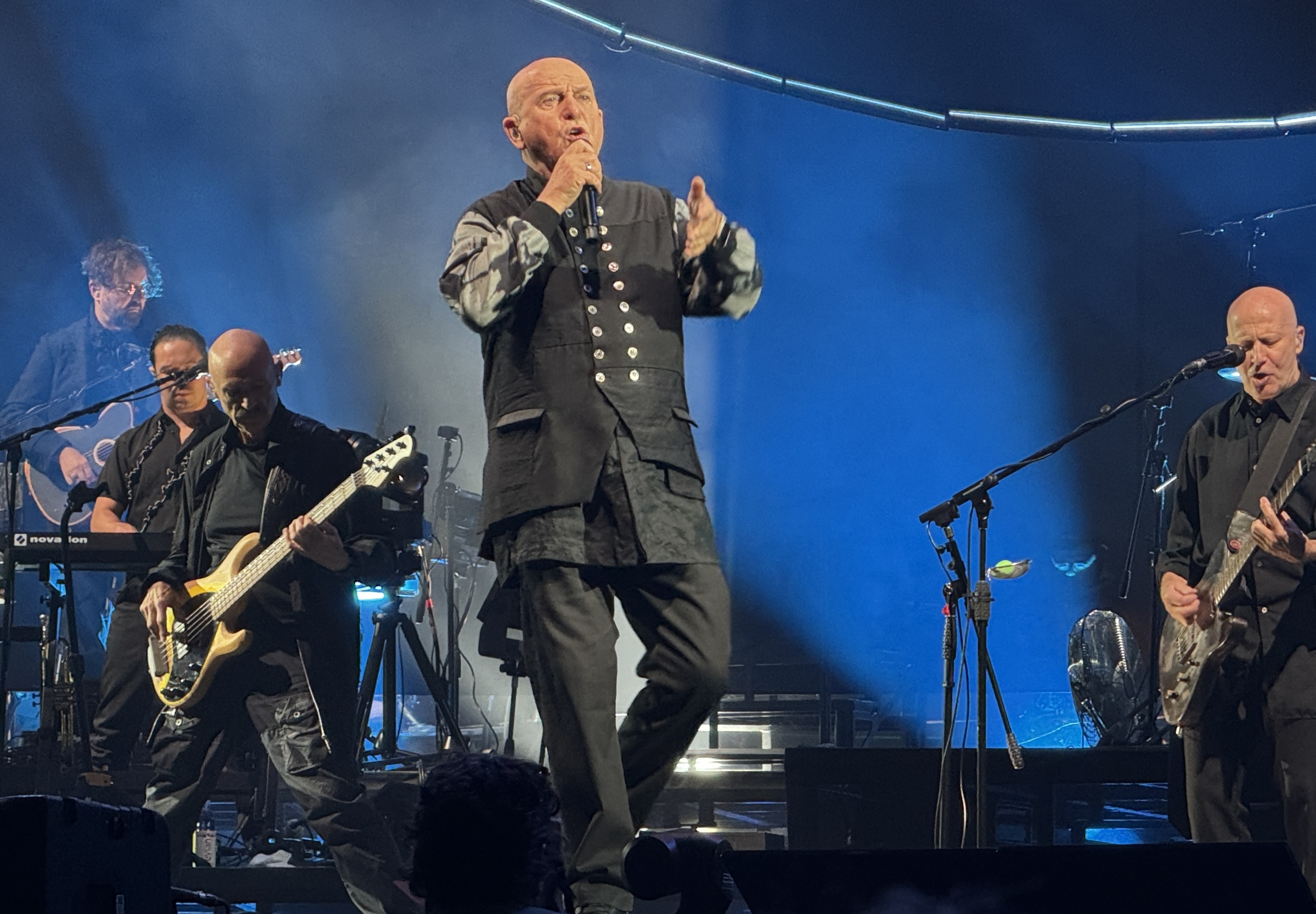 Peter Gabriel on stage singing with Tony Levin playing bass to his right and David Rhodes playing guitar to his left