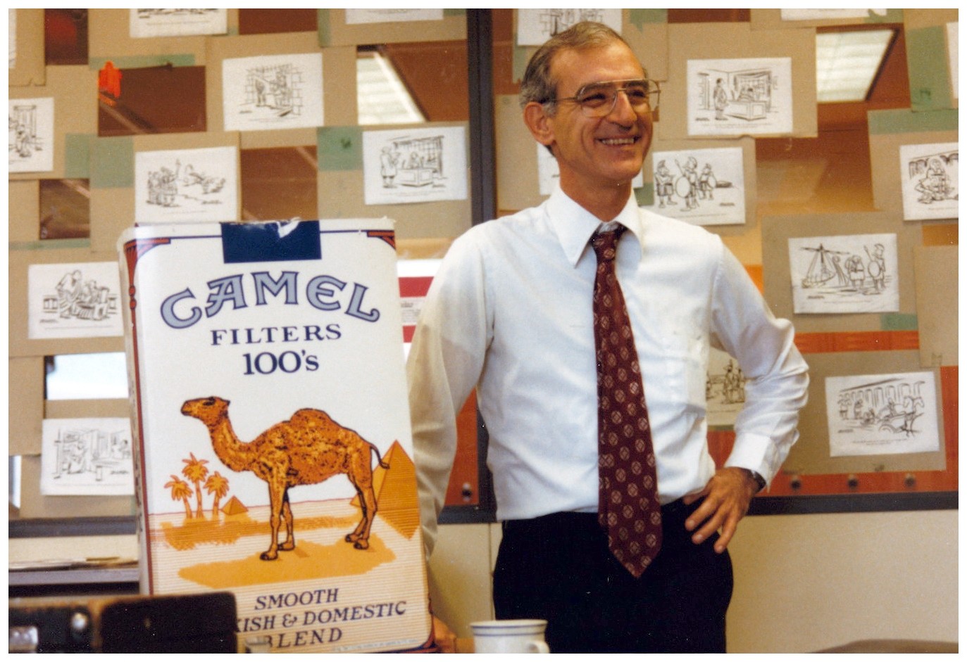 Saint Paul Central history teacher Richard Demers standing in his classroom with a giant pack of Camel cigarettes, smiling while wearing a white dress shirt, red tie and dark pants