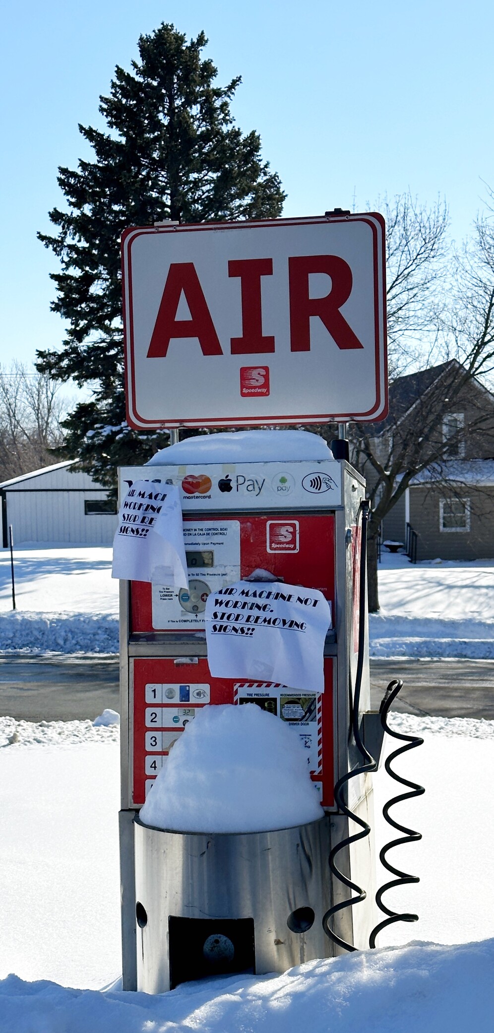 Tire air pump outside a gas station with paper sign taped to it that mentions it is broken and to stop removing the signs