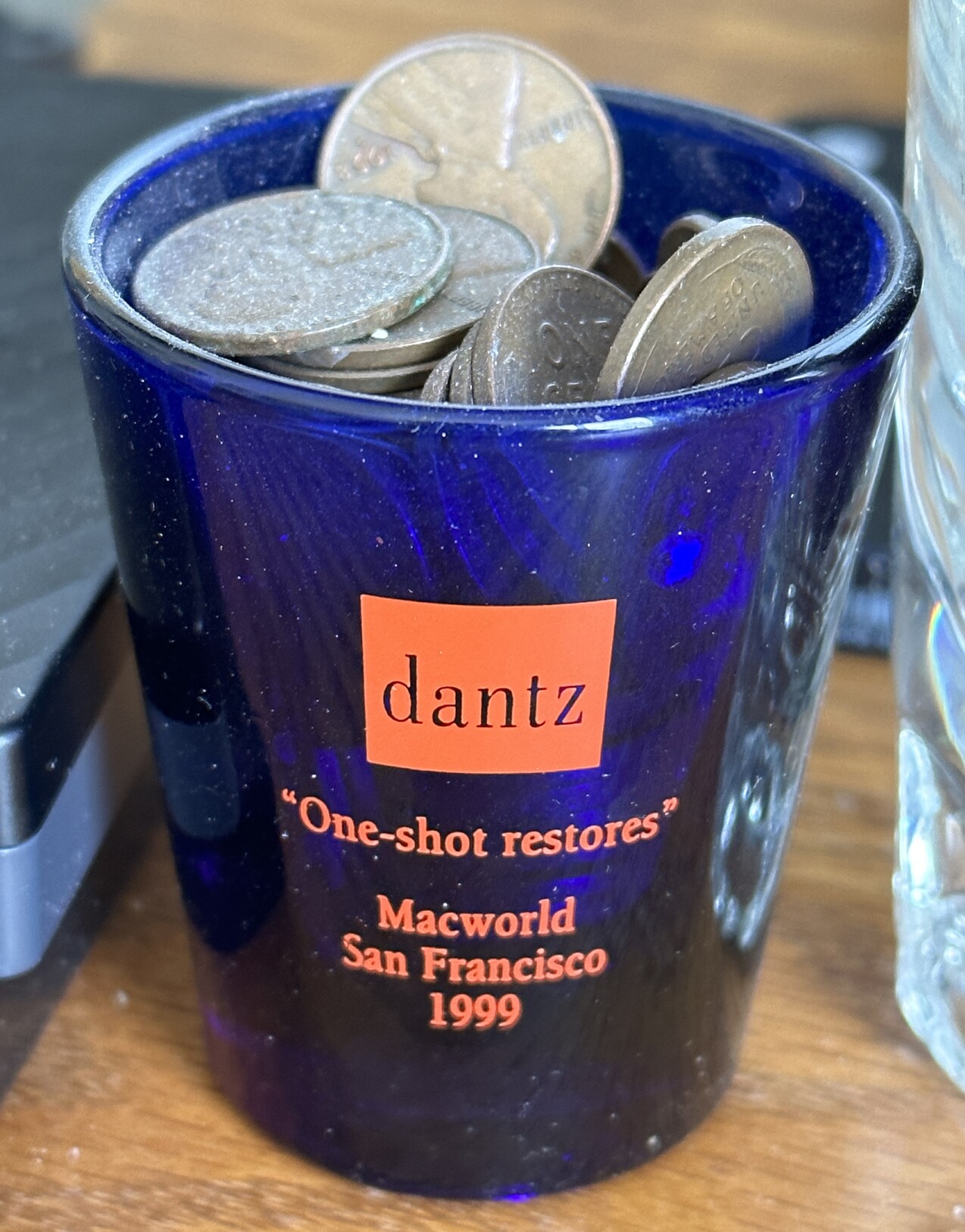 Blue Dantz shot glass from MacWorld San Francisco 1999 with red Dantz logo and lettering that says “One-shot restores” and is full of old, dusty pennies